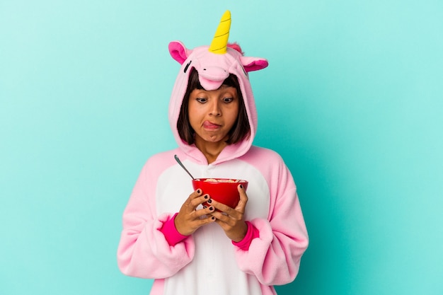 Joven mujer de raza mixta vistiendo un pijama de unicornio sosteniendo un cereal aislado sobre fondo azul.