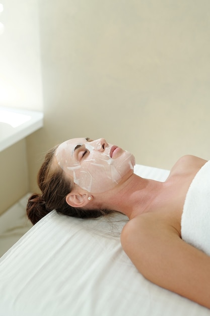 Joven mujer relajada con mascarilla en la cara disfrutando del procedimiento de cuidado de la piel en el lujoso salón de spa