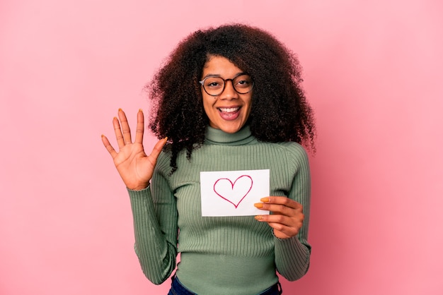 Joven mujer rizada afroamericana sosteniendo un símbolo de corazón en cartel sonriendo alegre mostrando el número cinco con los dedos.