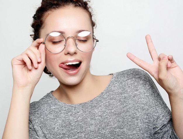 Joven mujer rizada con anteojos vestida de manera informal haciendo muecas a la cámara parpadeando sacando la lengua