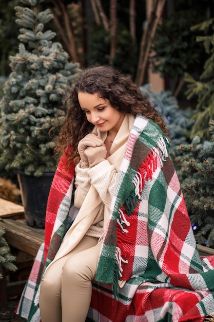 Una joven mujer rizada envuelta en una cálida tela escocesa en una celda se sienta esperando el nuevo año La niña en el fondo de verdes árboles de Navidad en la calle Estado de ánimo festivo