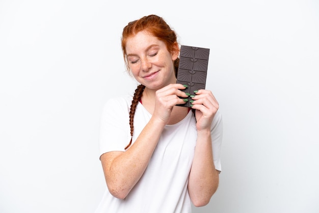 Joven mujer rojiza aislada de fondo blanco tomando una tableta de chocolate y feliz