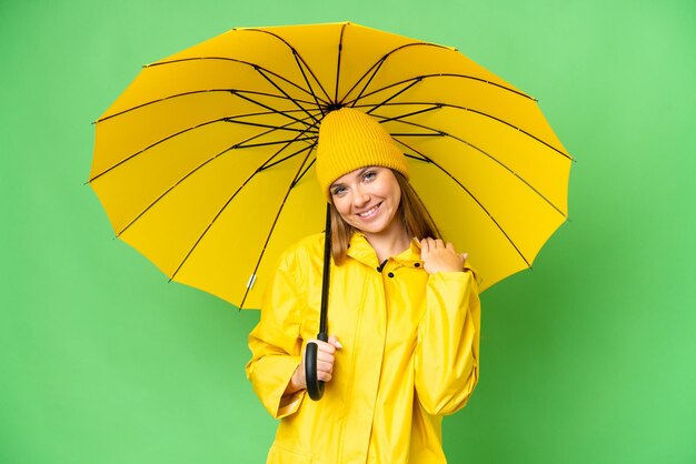 Joven mujer rubia con abrigo impermeable y paraguas sobre fondo aislado de clave de croma riendo