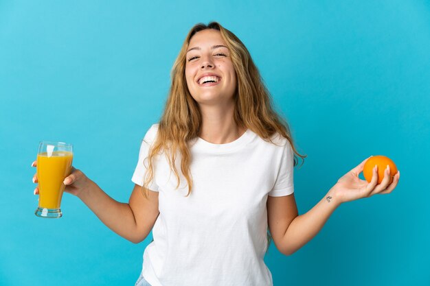 Joven mujer rubia aislada en la pared azul sosteniendo una naranja y un jugo de naranja