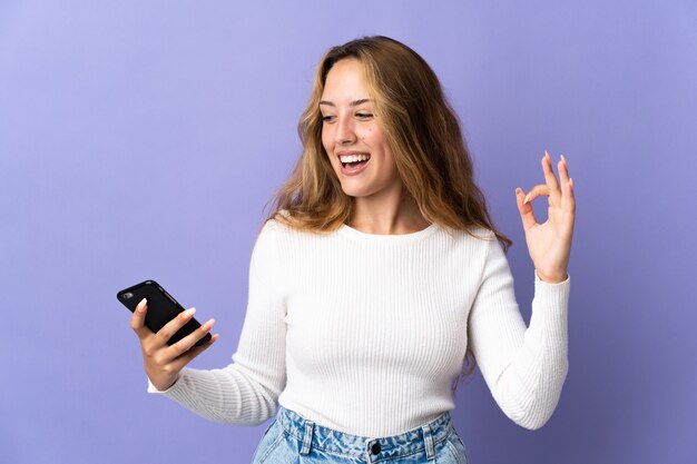 Joven mujer rubia aislada en la pared púrpura mediante teléfono móvil y haciendo el signo de OK