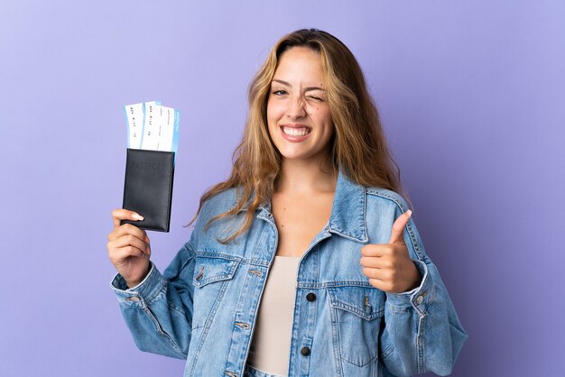 Joven mujer rubia aislada en la pared púrpura en vacaciones sosteniendo un pasaporte y avión con el pulgar hacia arriba