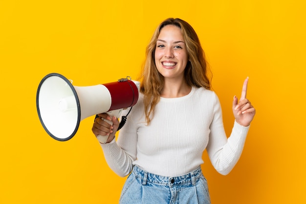 Joven mujer rubia aislada sobre fondo amarillo sosteniendo un megáfono y apuntando hacia una gran idea