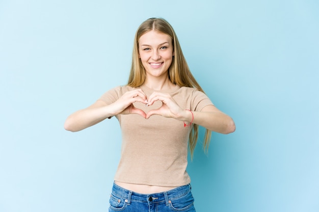 Joven mujer rubia aislada sobre fondo azul sonriendo y mostrando una forma de corazón con las manos.