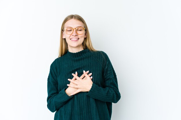 Joven mujer rubia aislada sobre fondo blanco riendo manteniendo las manos en el corazón, concepto de felicidad.