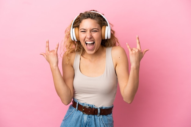 Joven mujer rubia aislada sobre fondo rosa escuchando música haciendo gesto de rock