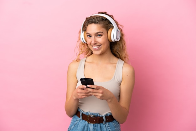 Joven mujer rubia aislada sobre fondo rosa escuchando música con un móvil y mirando al frente