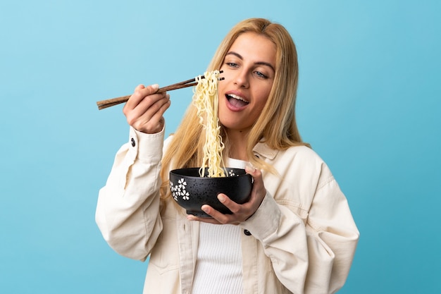 Joven mujer rubia aislada sosteniendo un plato de fideos con palillos y comiéndolo