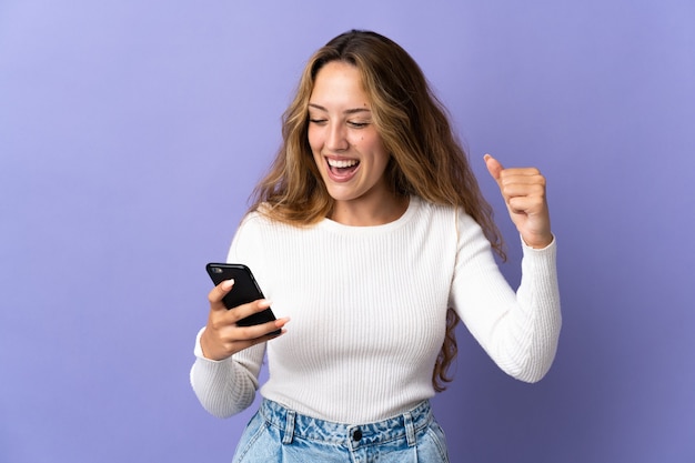 Joven mujer rubia aislada mediante teléfono móvil haciendo gesto de victoria