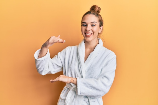 Joven mujer rubia con albornoz gesticulando con las manos mostrando un símbolo de medida de signo grande y grande sonriendo mirando el concepto de medición de la cámara