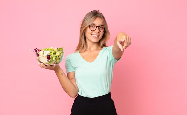 Joven mujer rubia apuntando a la cámara eligiéndote y sosteniendo una ensalada