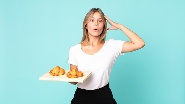 Joven mujer rubia con aspecto feliz, asombrado y sorprendido y sosteniendo una bandeja de croissant