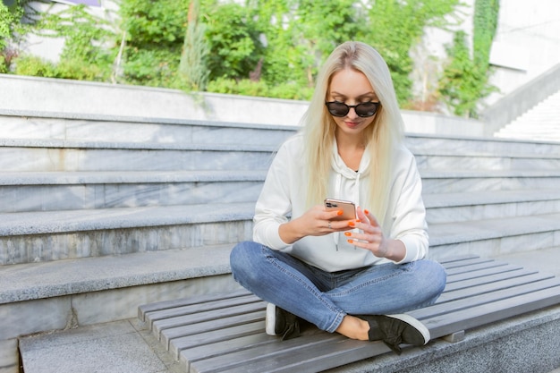 Joven mujer rubia atractiva con gafas de sol usando un teléfono inteligente mientras se sienta en un banco en el parque de la ciudad