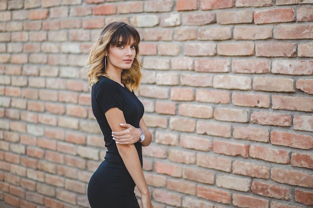 Joven mujer rubia atractiva con un vestido negro posando frente a una pared de ladrillos