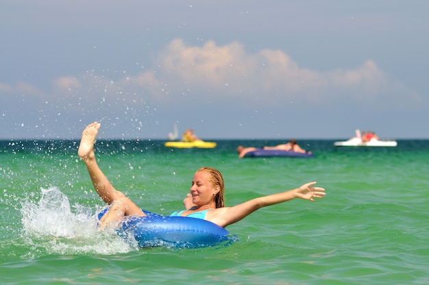 Joven mujer rubia en bikini azul montando en círculo de natación en el mar y sonriendo en un día soleado de verano. Concepto de felicidad, vacaciones y libertad