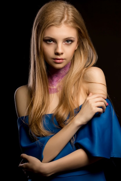 Joven mujer rubia con cabello largo y exuberante y vestido azul con hombros desnudos posando en el estudio