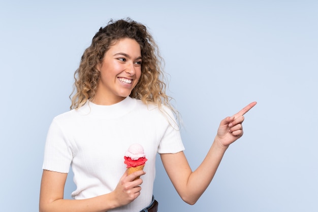 Joven mujer rubia con cabello rizado sosteniendo un helado de cucurucho aislado en la pared azul apuntando hacia el lado para presentar un producto