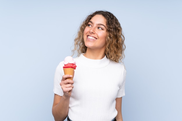 Joven mujer rubia con cabello rizado sosteniendo un helado de cucurucho aislado en la pared azul mirando hacia arriba mientras sonríe