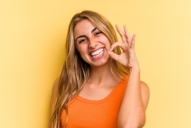 Joven mujer rubia caucásica aislada sobre fondo amarillo alegre y confiado mostrando gesto ok.