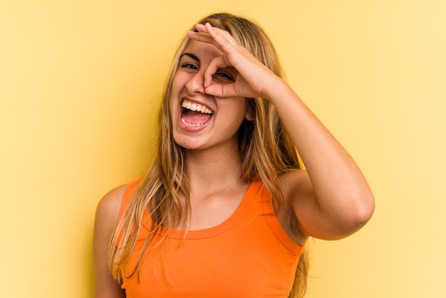 Joven mujer rubia caucásica aislada sobre fondo amarillo emocionado manteniendo el gesto ok en el ojo.