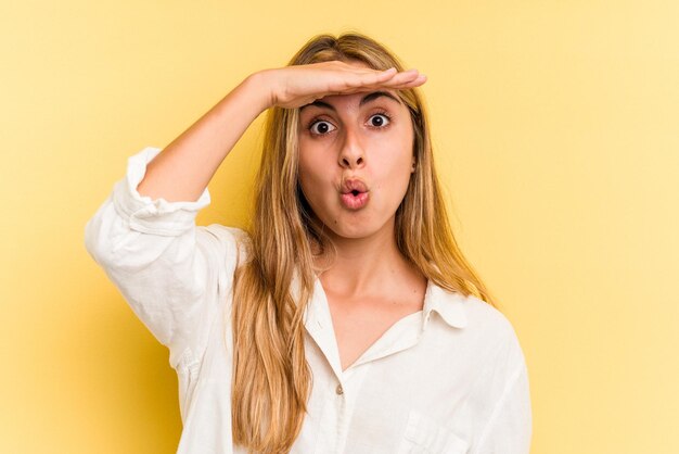 Joven mujer rubia caucásica aislada sobre fondo amarillo mirando lejos manteniendo la mano en la frente.