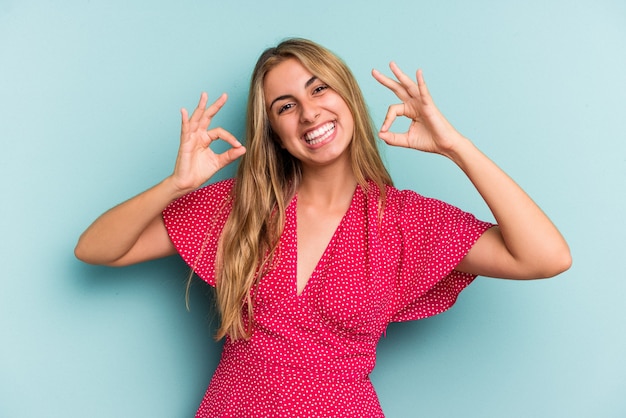 Joven mujer rubia caucásica aislada sobre fondo azul alegre y confiado mostrando gesto ok.