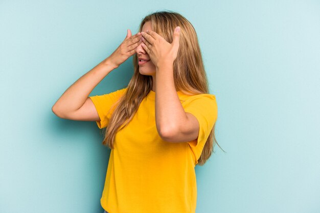 Joven mujer rubia caucásica aislada sobre fondo azul miedo cubrirse los ojos con las manos.