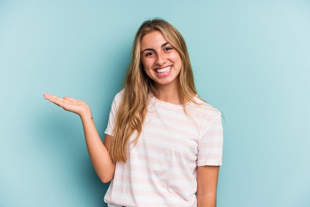 Joven mujer rubia caucásica aislada sobre fondo azul mostrando un espacio de copia en una palma y sosteniendo otra mano en la cintura.