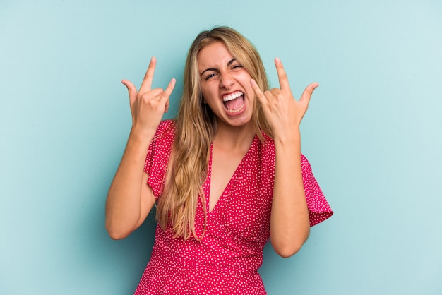 Joven mujer rubia caucásica aislada sobre fondo azul mostrando un gesto de cuernos como un concepto de revolución.