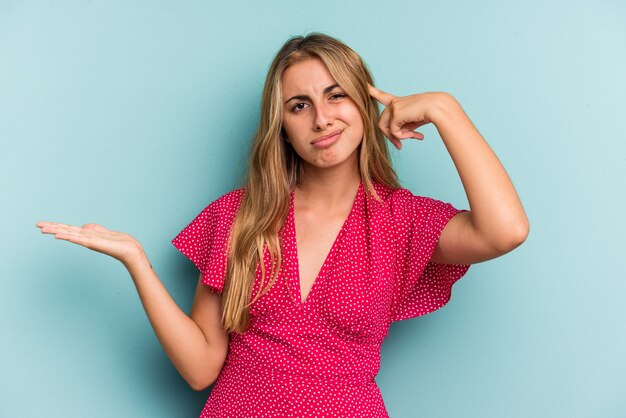 Joven mujer rubia caucásica aislada sobre fondo azul mostrando un gesto de decepción con el dedo índice.