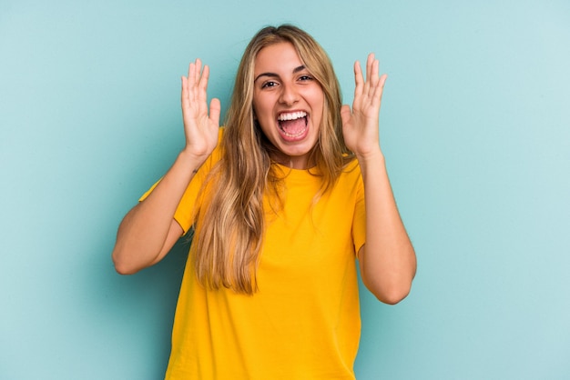 Joven mujer rubia caucásica aislada sobre fondo azul recibiendo una agradable sorpresa, emocionada y levantando las manos.