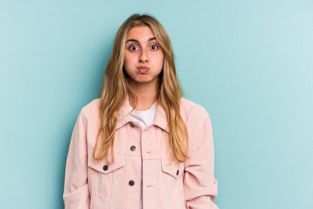 Joven mujer rubia caucásica aislada sobre fondo azul sopla mejillas, tiene expresión cansada. Concepto de expresión facial.