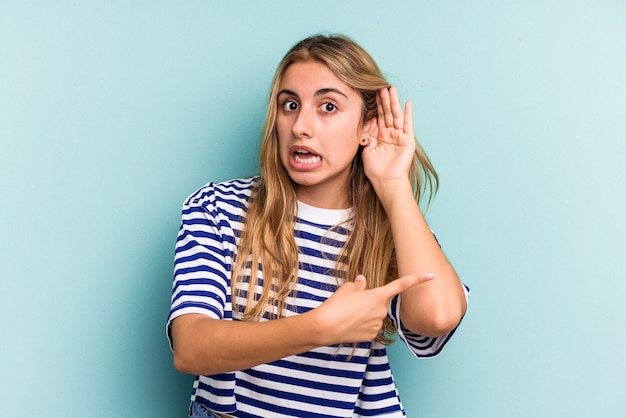 Joven mujer rubia caucásica aislada sobre fondo azul tratando de escuchar un chisme.