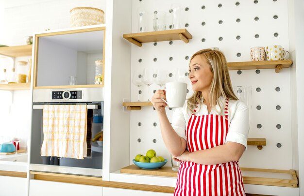 Joven mujer rubia caucásica con delantal de rayas rojas sosteniendo la taza mirando al lado sonriendo en la cocina