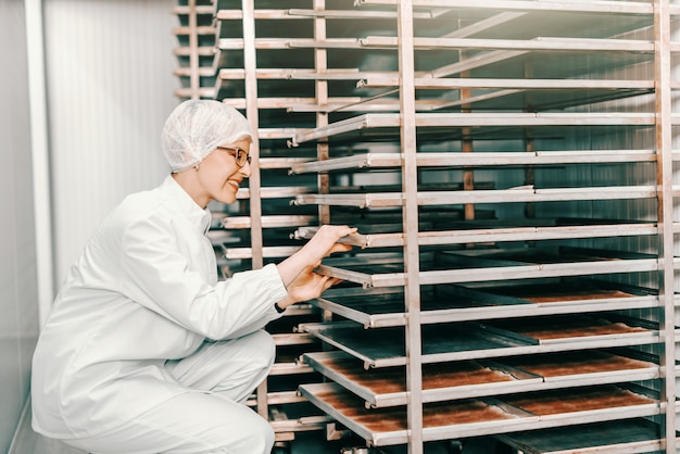 Joven mujer rubia caucásica empleado en uniforme estéril poniendo bandejas en el estante mientras se agacha en la fábrica de alimentos.