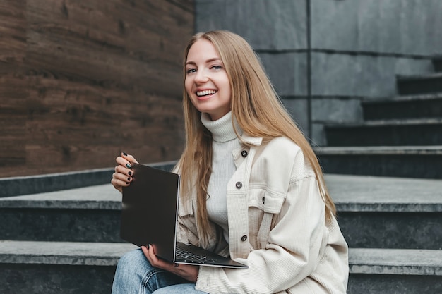 Joven mujer rubia caucásica especialista en TI sentado en las escaleras cerca del edificio de oficinas con un portátil sonriendo