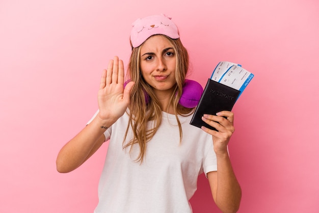 Joven mujer rubia caucásica sosteniendo un pasaporte y boletos para viajar aislado sobre fondo rosa de pie con la mano extendida mostrando la señal de stop, impidiéndote.