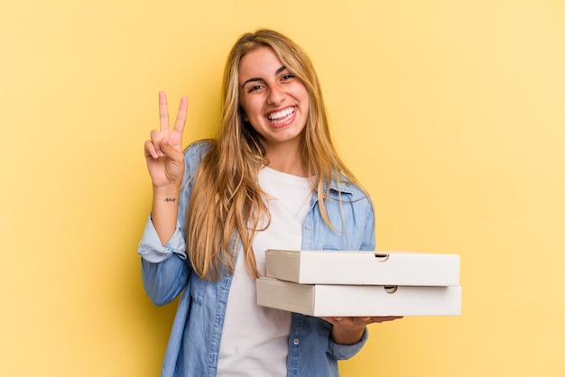 Joven mujer rubia caucásica sosteniendo pizzas aisladas sobre fondo amarillo mostrando el número dos con los dedos.