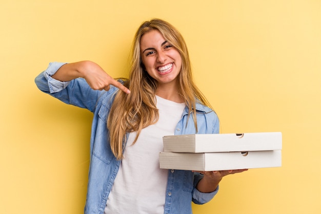 Joven mujer rubia caucásica sosteniendo pizzas aisladas sobre fondo amarillo persona apuntando con la mano a un espacio de copia de camisa, orgulloso y seguro
