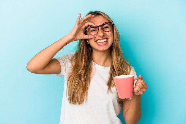 Joven mujer rubia caucásica sosteniendo una taza aislada sobre fondo azul emocionado manteniendo el gesto ok en el ojo.