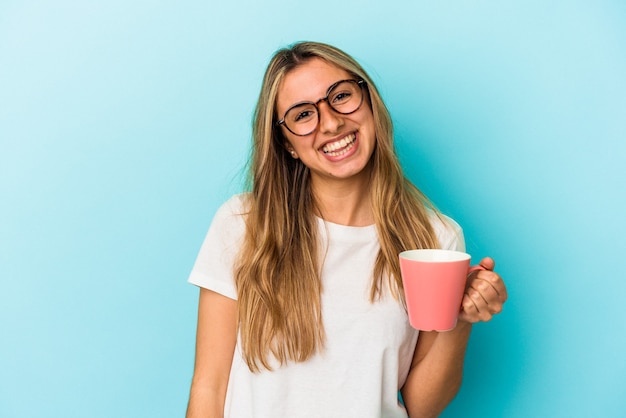 Joven mujer rubia caucásica sosteniendo una taza aislada sobre fondo azul feliz, sonriente y alegre.