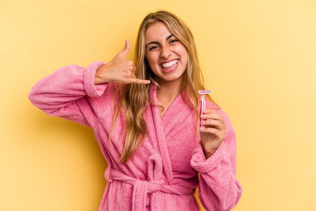 Joven mujer rubia caucásica vistiendo albornoz sosteniendo cuchillas aisladas sobre fondo amarillo mostrando un gesto de llamada de teléfono móvil con los dedos.