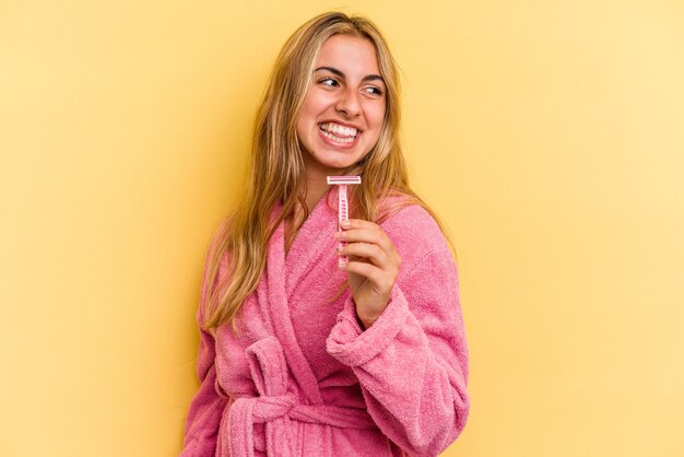 Joven mujer rubia caucásica vistiendo albornoz sosteniendo cuchillas aisladas sobre fondo amarillo se ve a un lado sonriente, alegre y agradable.