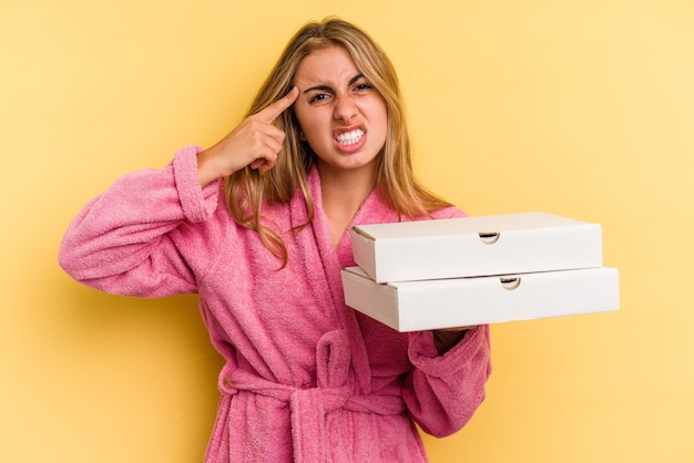 Joven mujer rubia caucásica vistiendo albornoz sosteniendo pizzas aisladas sobre fondo amarillo mostrando un gesto de decepción con el dedo índice.