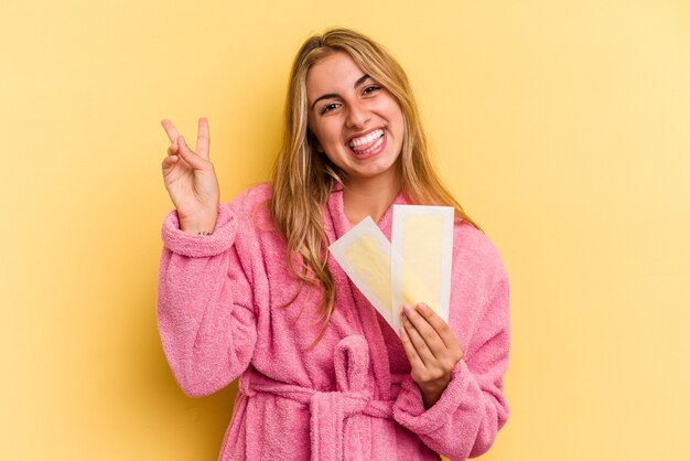 Joven mujer rubia caucásica vistiendo bata de baño sosteniendo bandas depilatorias aisladas sobre fondo amarillo alegre y despreocupado mostrando un símbolo de paz con los dedos.