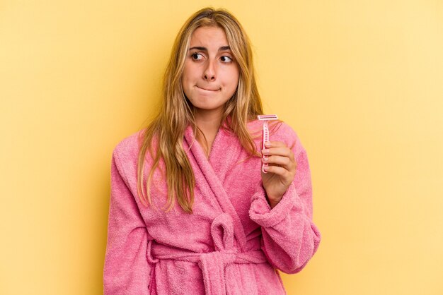 Joven mujer rubia caucásica vistiendo bata de baño sosteniendo cuchillas aisladas sobre fondo amarillo confundida, se siente dudosa e insegura.